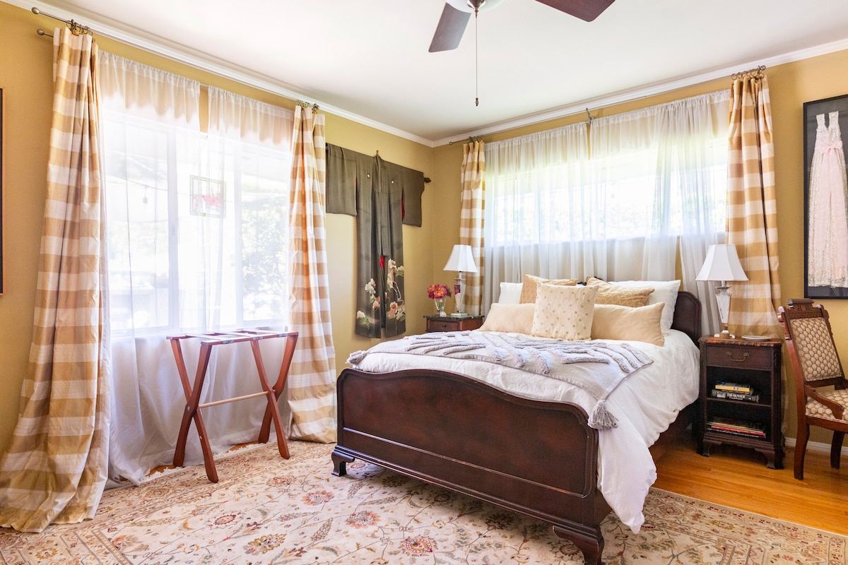 The bedroom features a spacious bed adorned with soft linens and decorative pillows. Large windows dressed with sheer and checkered curtains allow natural light to fill the room. A tasteful area rug covers the floor, and a bedside table with a lamp complements the decor.