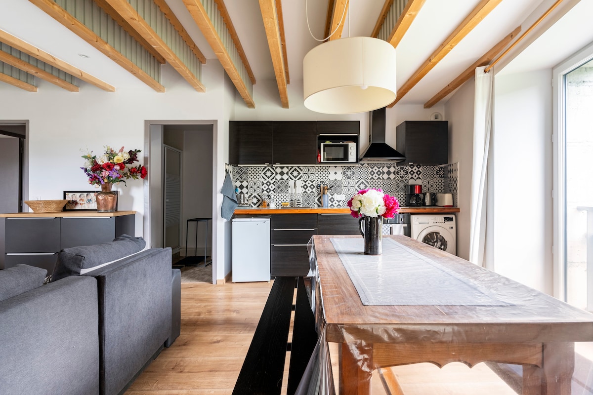 A modern kitchen is visible, featuring wooden beams overhead and a mix of sleek cabinetry with patterned tiles. An open layout connects the kitchen to a dining area with a wooden table, complemented by a cozy seating arrangement. Fresh flowers are arranged on the table.