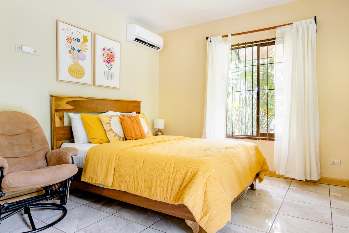 A bright bedroom features a spacious bed, accented with yellow bedding and decorative pillows. A wooden rocking chair is positioned nearby, and large windows adorned with white curtains allow natural light to enter the room. Artwork on the wall adds a touch of color.