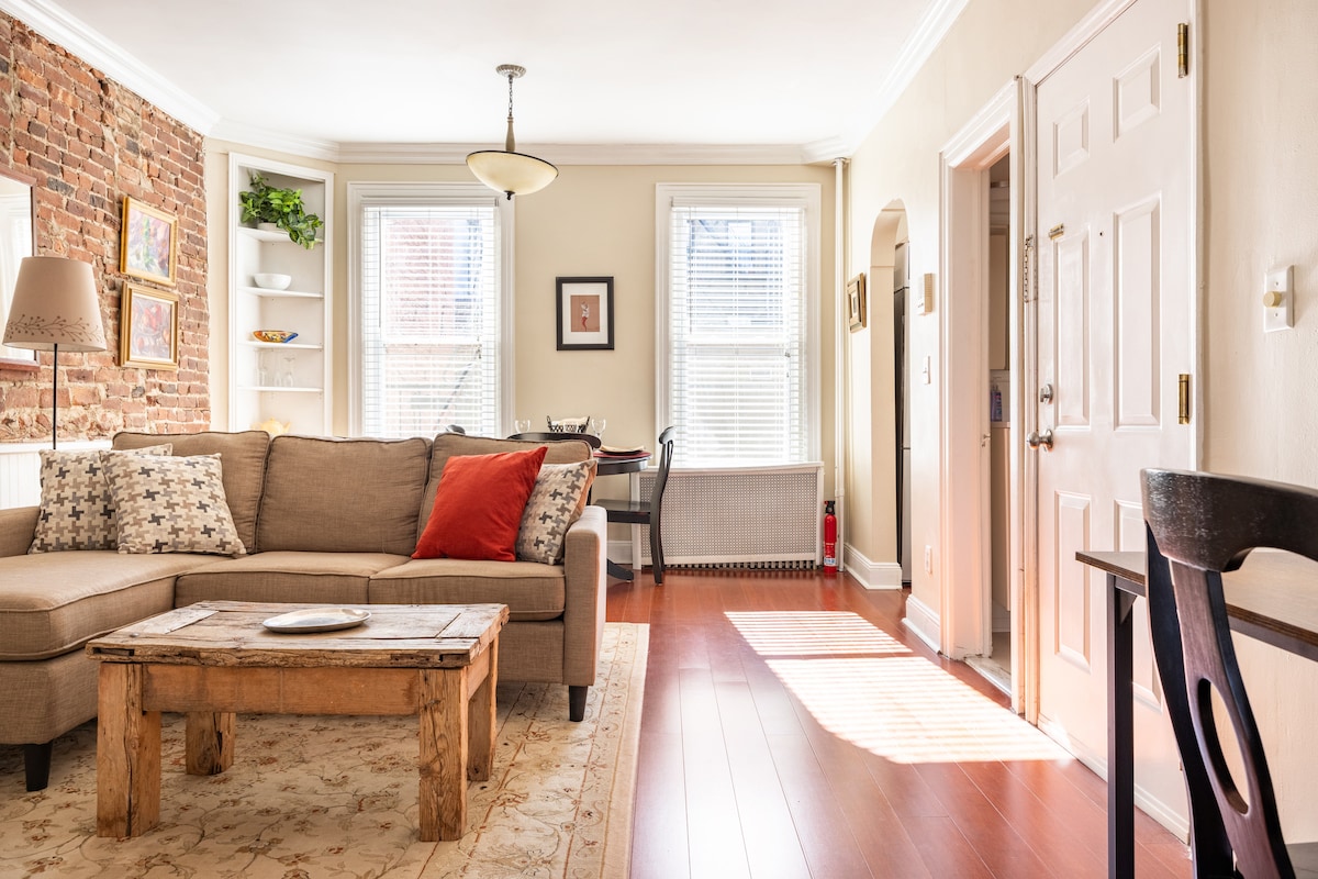 A cozy living area features a sectional sofa with decorative pillows, and a wooden coffee table sits in front. Bright light filters through large windows, illuminating the warm wooden floors. A dining table for two is visible, along with a decorative shelf showcasing greenery.