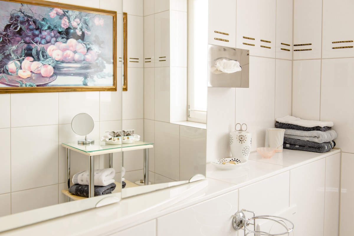 The well-appointed bathroom features white tile walls, providing a clean and bright atmosphere. A large mirror reflects the space, while decorative elements include a fruit-themed painting and neatly arranged towels. A small cart holds toiletries, adding to the room's functional charm.