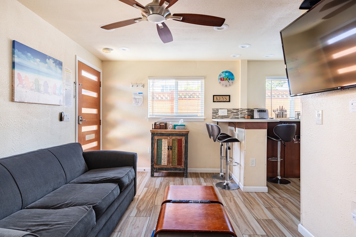 A cozy living area features a dark gray sofa paired with a wooden coffee table. A small bar with black seating is seen in the background, alongside a wall-mounted television. Natural light enters through two windows, enhancing the warm atmosphere of the space.