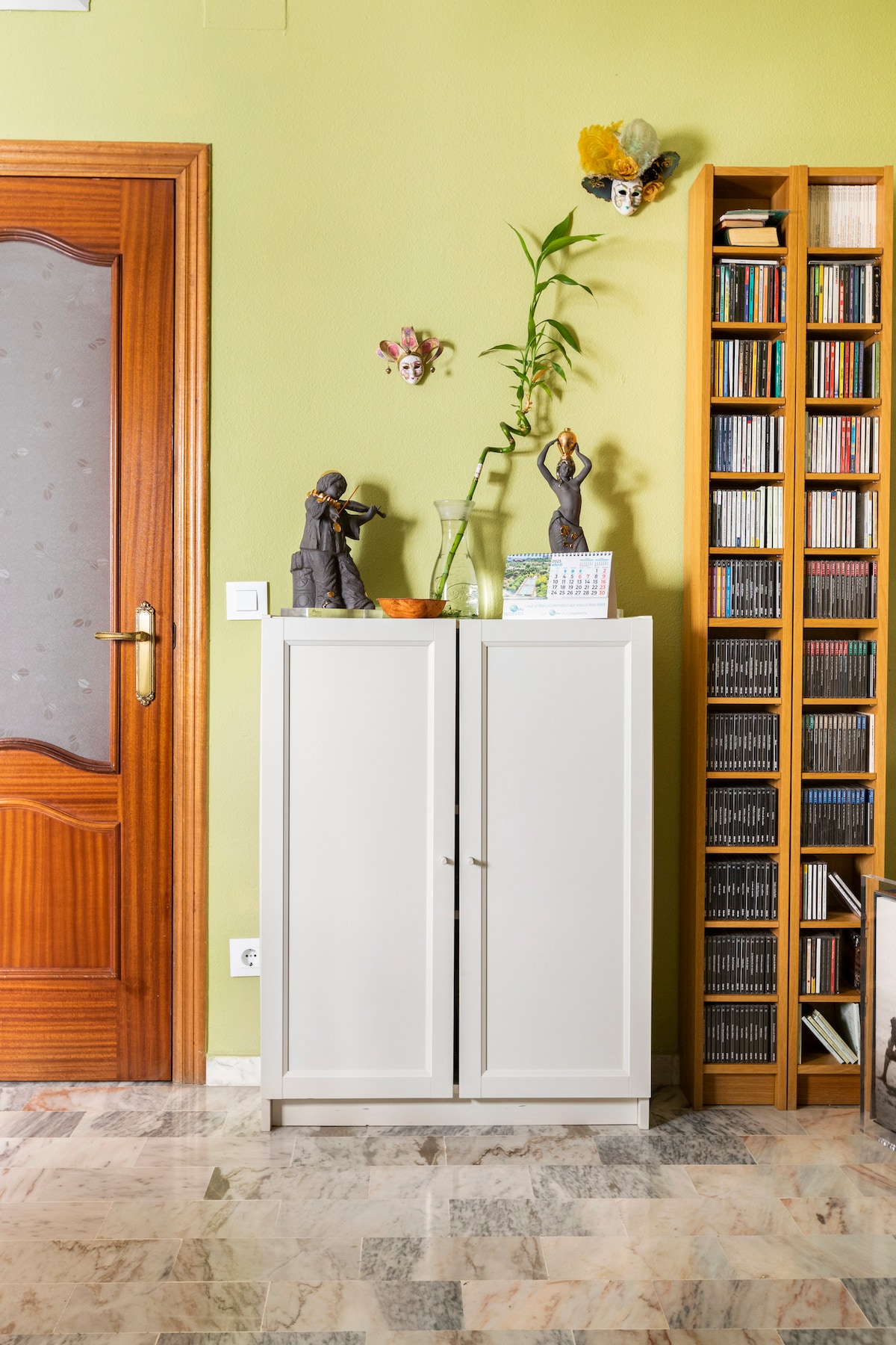 A minimalist storage cabinet in white stands against a green wall, topped with decorative items. A tall plant and a glass vase add a touch of nature. A wooden bookshelf filled with CDs is positioned nearby, providing an organized and inviting look.