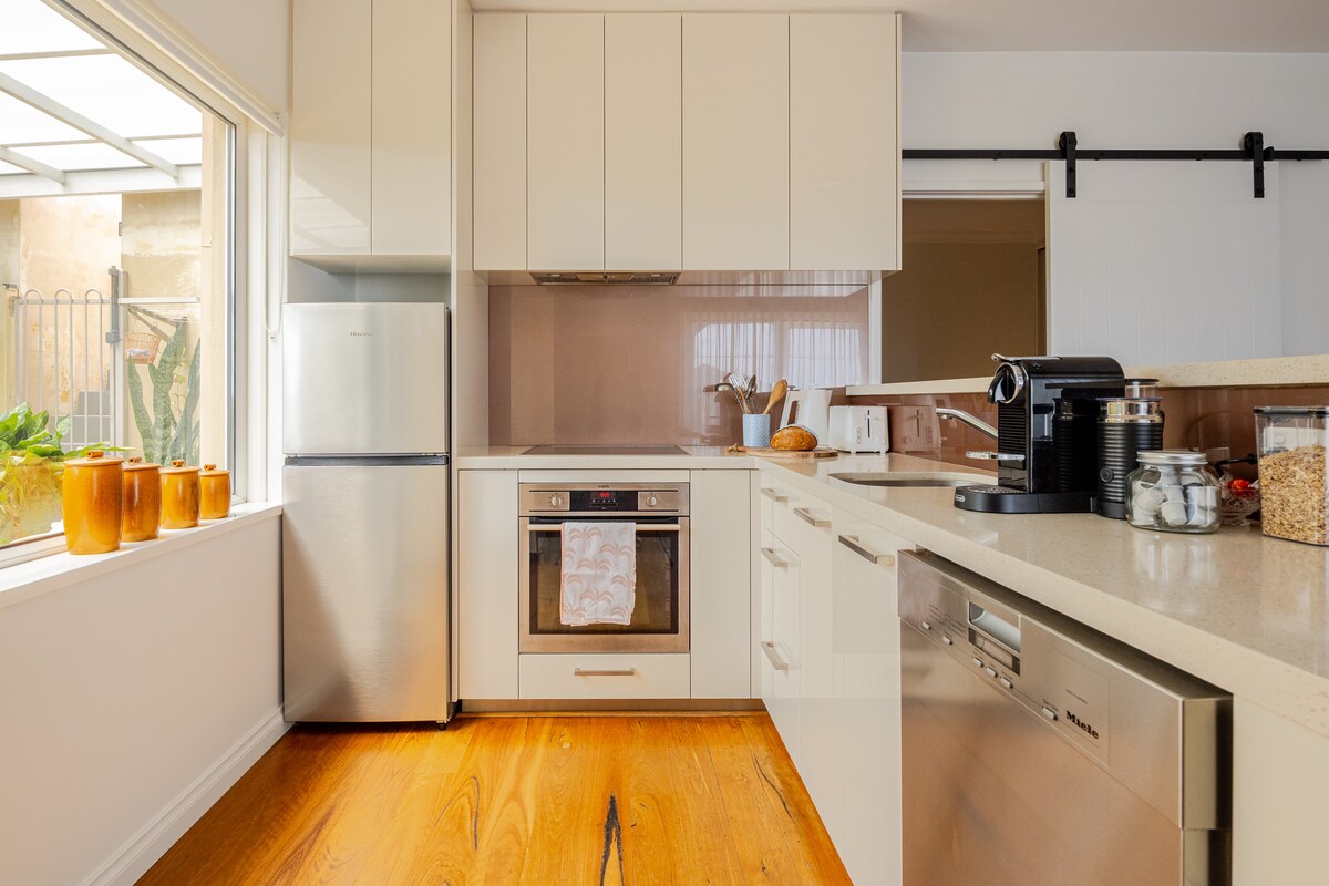 The kitchen features modern cabinetry and appliances, including a full-sized refrigerator, built-in oven, and dishwasher. A Nespresso machine is also provided. Natural light fills the space through a large window, highlighting the sleek countertop and wooden flooring.