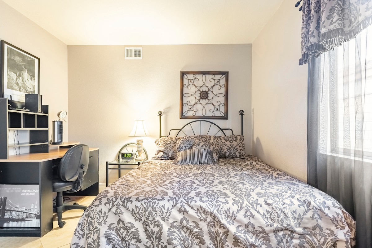 A well-appointed room features a queen bed adorned with a patterned comforter. A desk with a chair sits adjacent, providing a workspace. Soft lighting is provided by a bedside lamp, while natural light filters through a sheer curtain at the window.