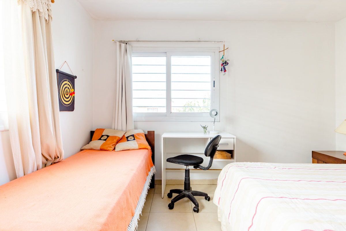 A bright bedroom features two beds, one with an orange coverlet and the other with a light multicolored duvet. A desk with a chair is positioned by a window with horizontal slats, allowing natural light to illuminate the space.
