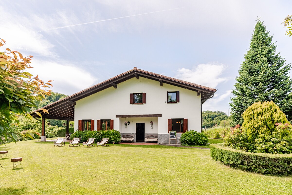 The house features a spacious two-story façade with a porch, framed by well-maintained greenery. Lounge chairs are set on the lawn, which is neatly trimmed and surrounded by various shrubs and trees, enhancing the natural landscape.