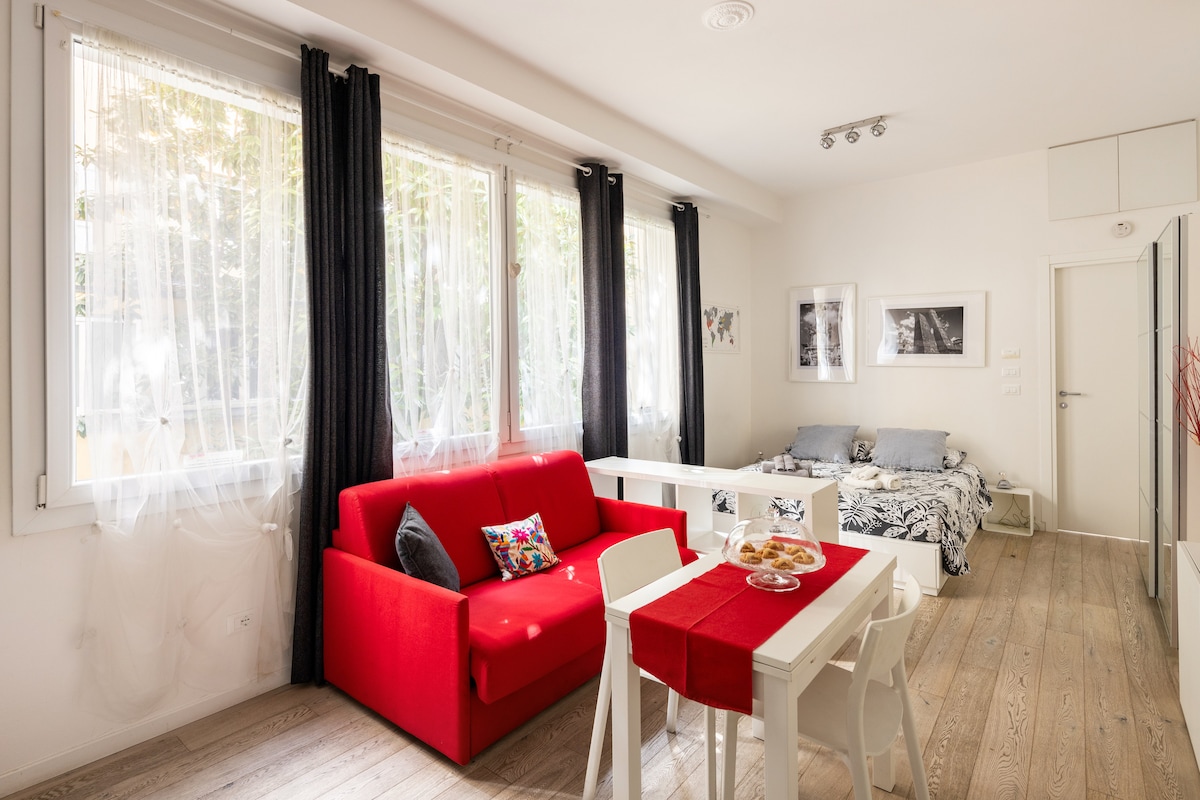 A bright living area features a red sofa and a white dining table with chairs. Sunlight streams through large windows, draped with sheer curtains, illuminating the space. A bed with patterned linens is positioned nearby. Art photographs adorn the walls.