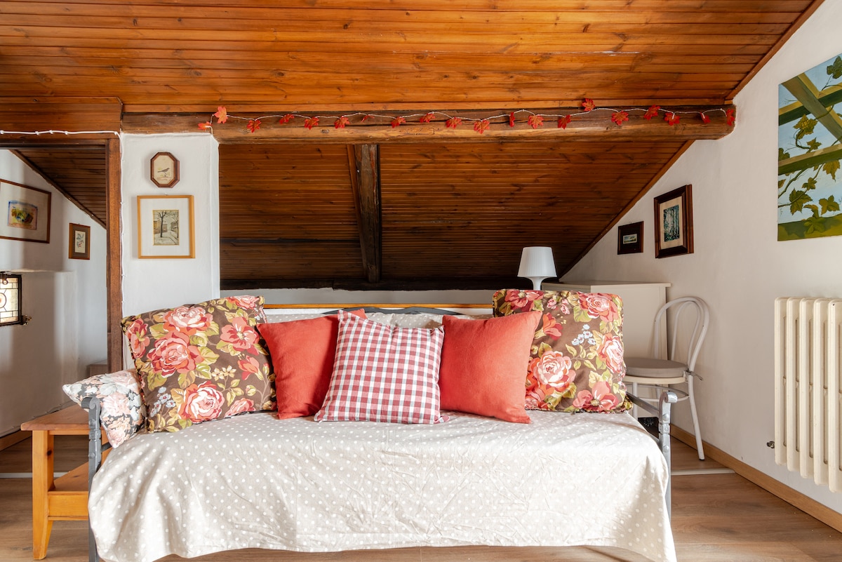 A cozy seating area features a daybed adorned with floral and checkered pillows. The warm wooden ceiling adds to the inviting ambiance, while framed artworks are displayed on the walls. A subtle light source illuminates the space, enhancing its charm.