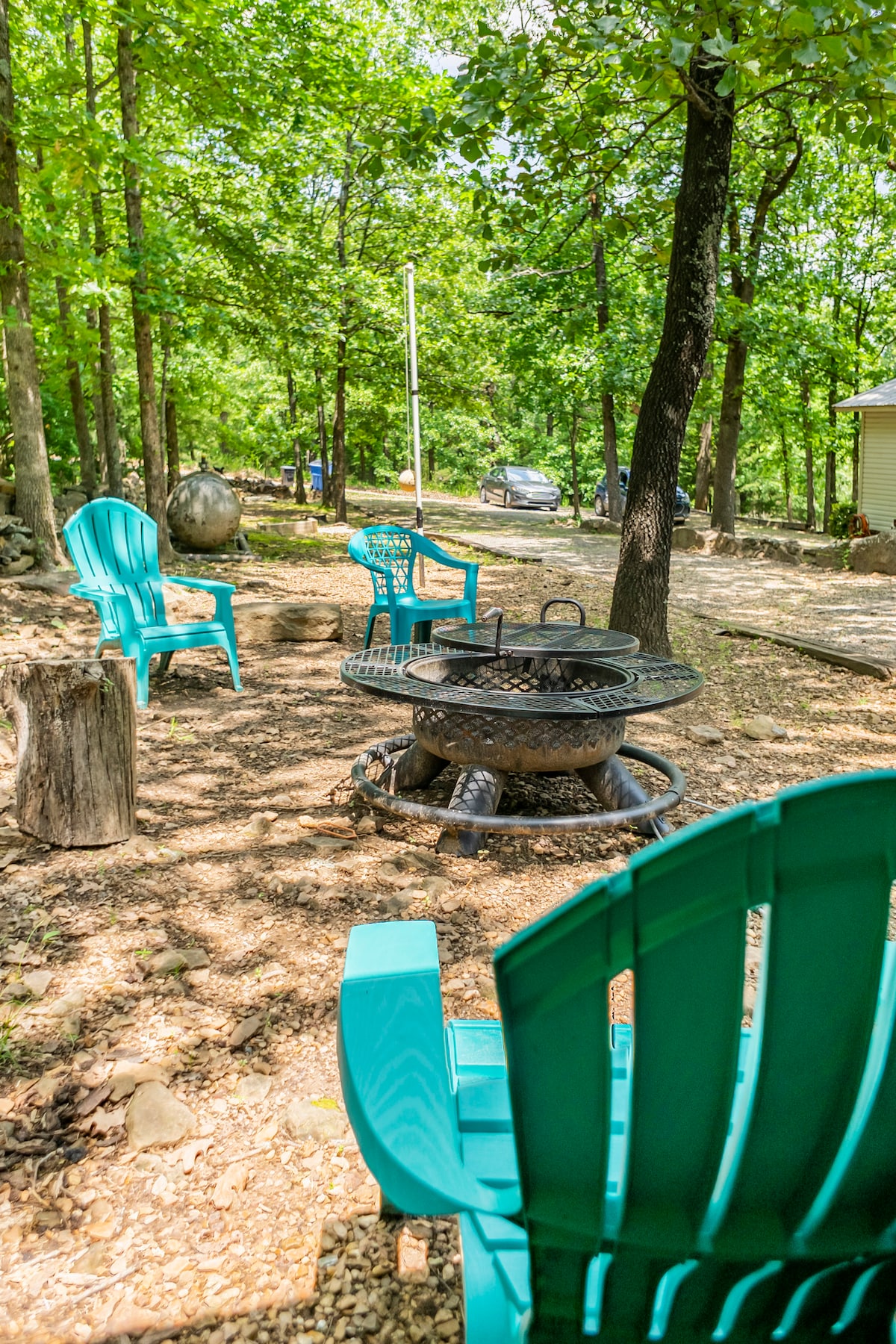 A fire pit surrounded by several teal adirondack chairs is situated on a natural ground area. Green trees provide shade in the background, enhancing a relaxed outdoor space for gatherings. A vehicle is visible in the distance along the gravel path.