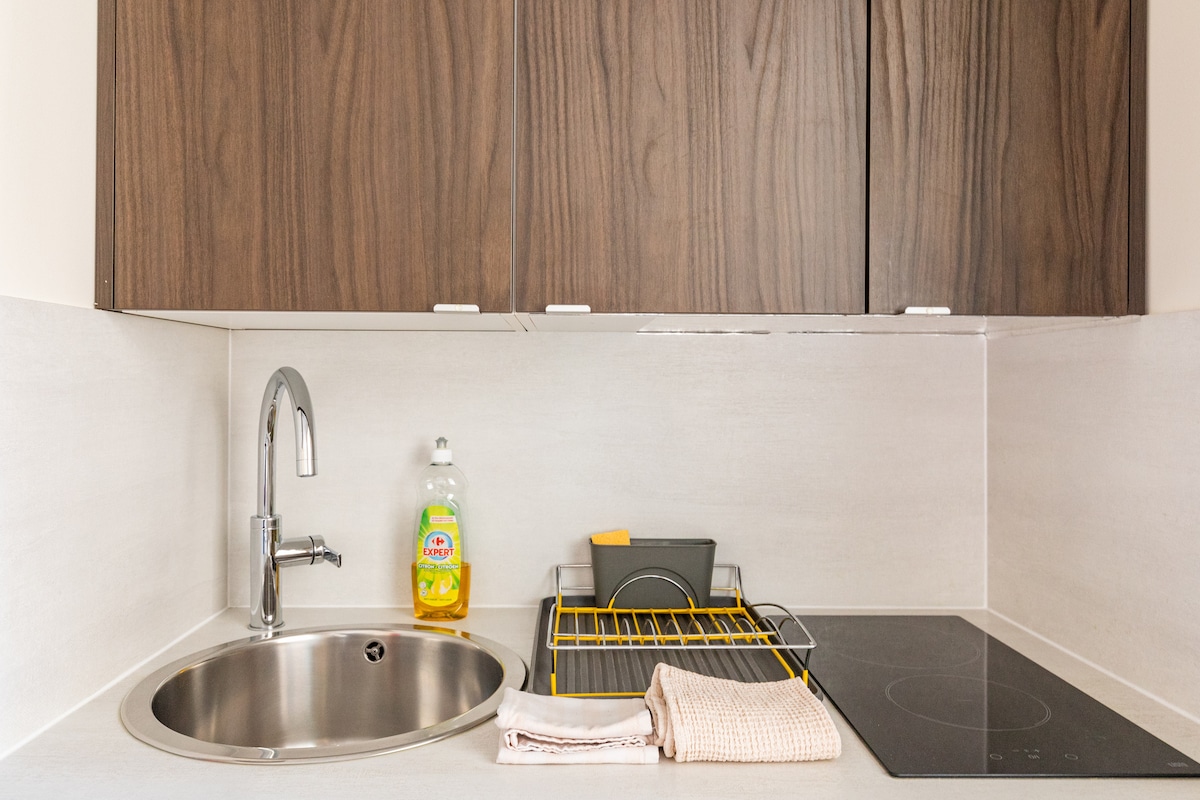 A compact kitchen area showcases a stainless steel sink with a modern faucet. A bottle of dish soap is placed beside a drying rack fitted with clean dishes. An electric cooktop is visible, alongside neatly arranged kitchen towels, adding to the functionality of the space.