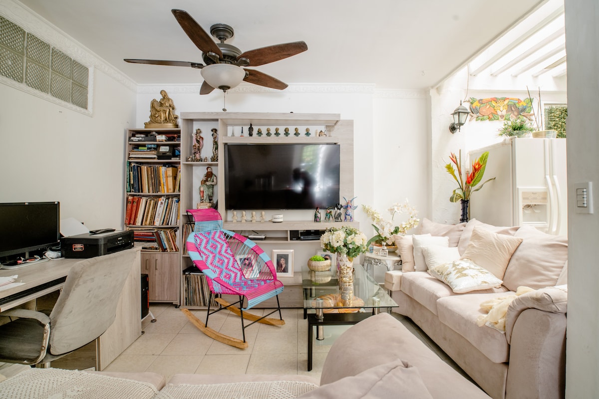 A living area contains a large wall-mounted television and a light-colored sectional sofa. A rocking chair with a colorful blanket is featured, along with a glass coffee table displaying decorative items. Shelves are lined with books, and a small indoor garden is visible.