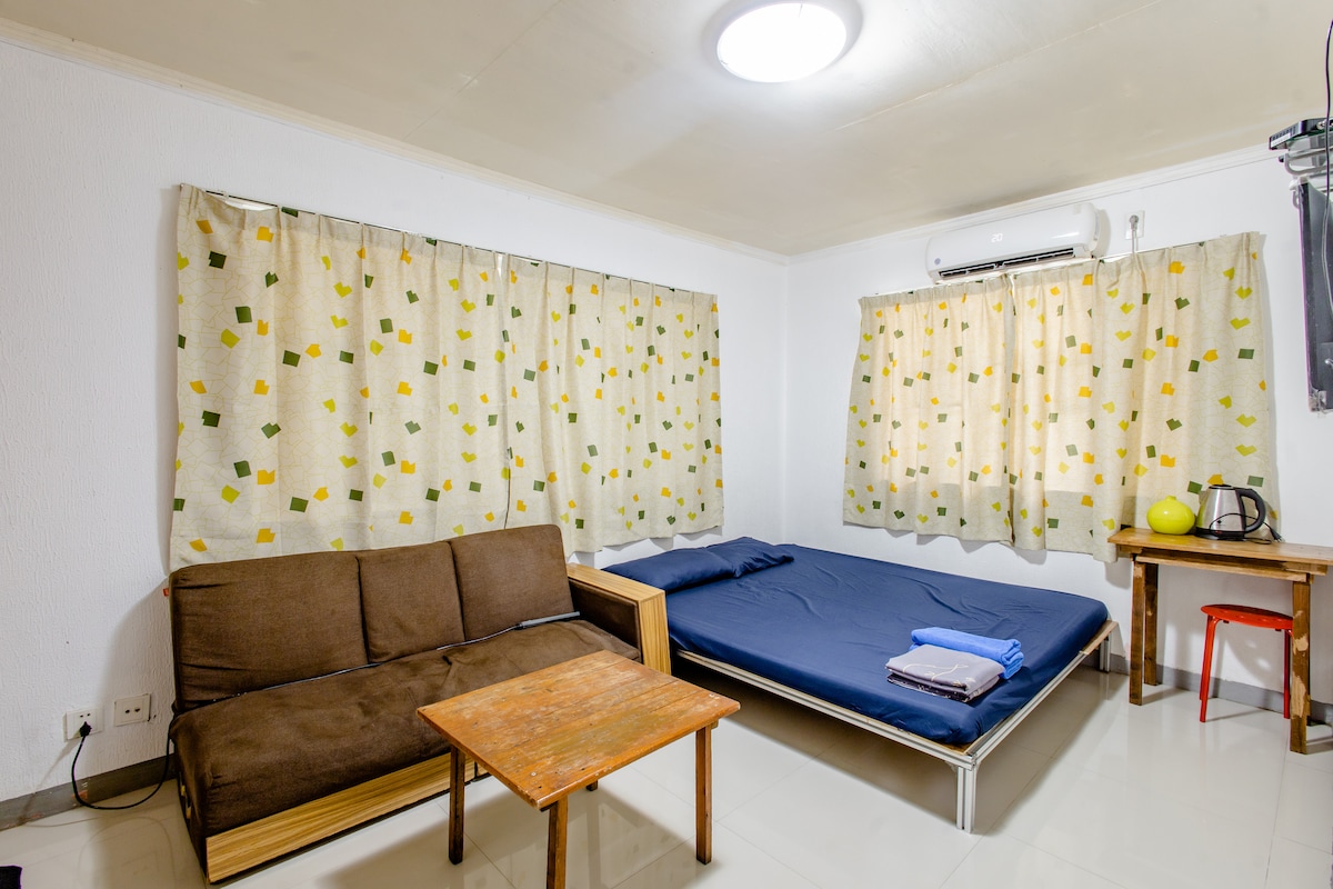 A minimalistic room is equipped with a spacious bed made up with blue bedding and neatly folded towels. A brown sofa is positioned opposite a wooden table, accompanied by a kettle and a bowl of fruit. Natural light filters through patterned curtains.