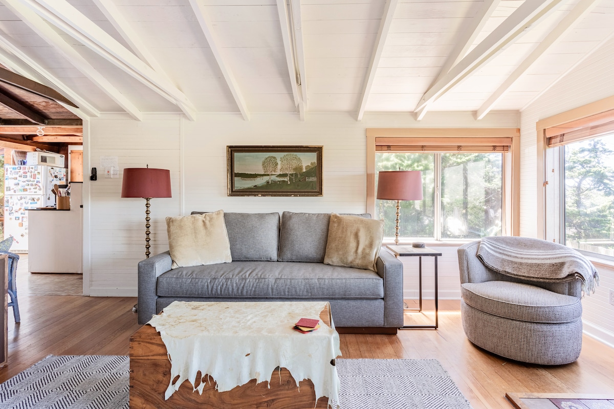 A spacious living area features a gray sofa adorned with plush pillows, complemented by a round accent chair. Natural light fills the room through large windows, highlighting a wooden coffee table with a decorative piece. Wooden beams create an inviting ambiance.