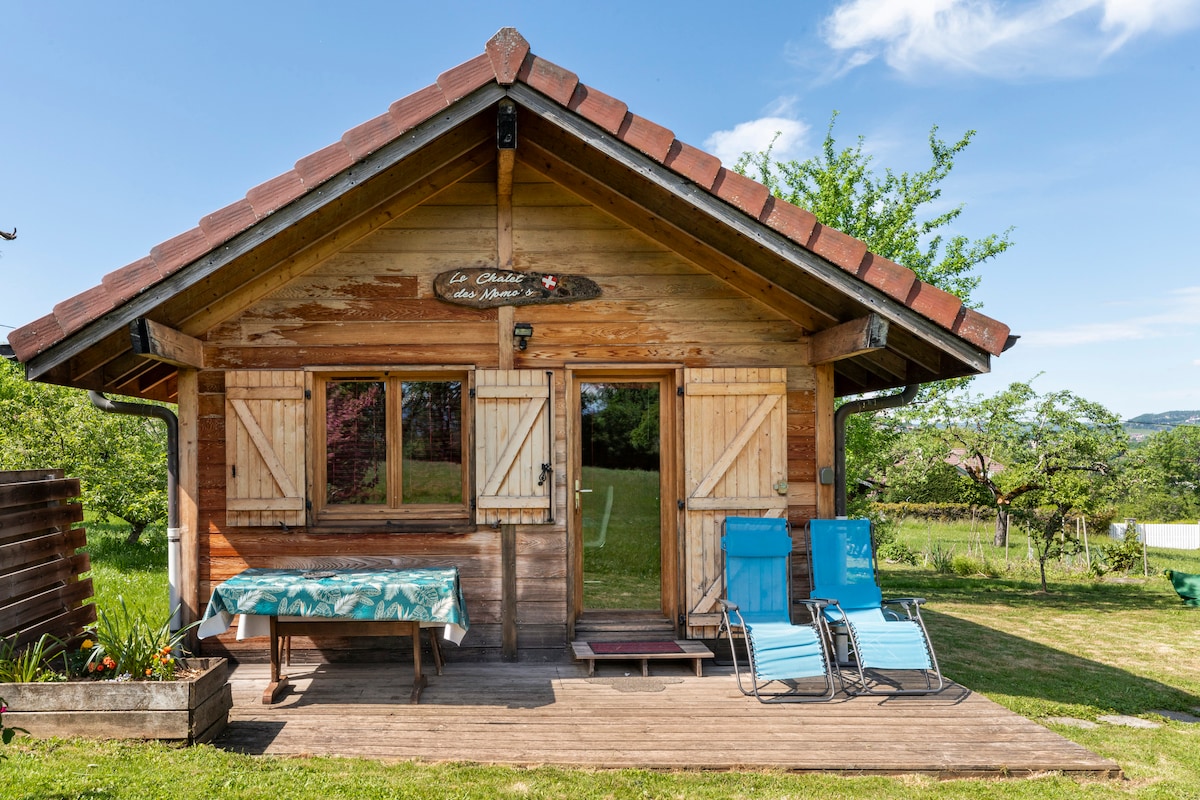 A charming wooden chalet is shown from the front, featuring a sloped roof and inviting entryway. A small table with a patterned tablecloth is positioned outside, accompanied by two blue lounge chairs on the grass. Lush greenery surrounds the building, enhancing the natural setting.