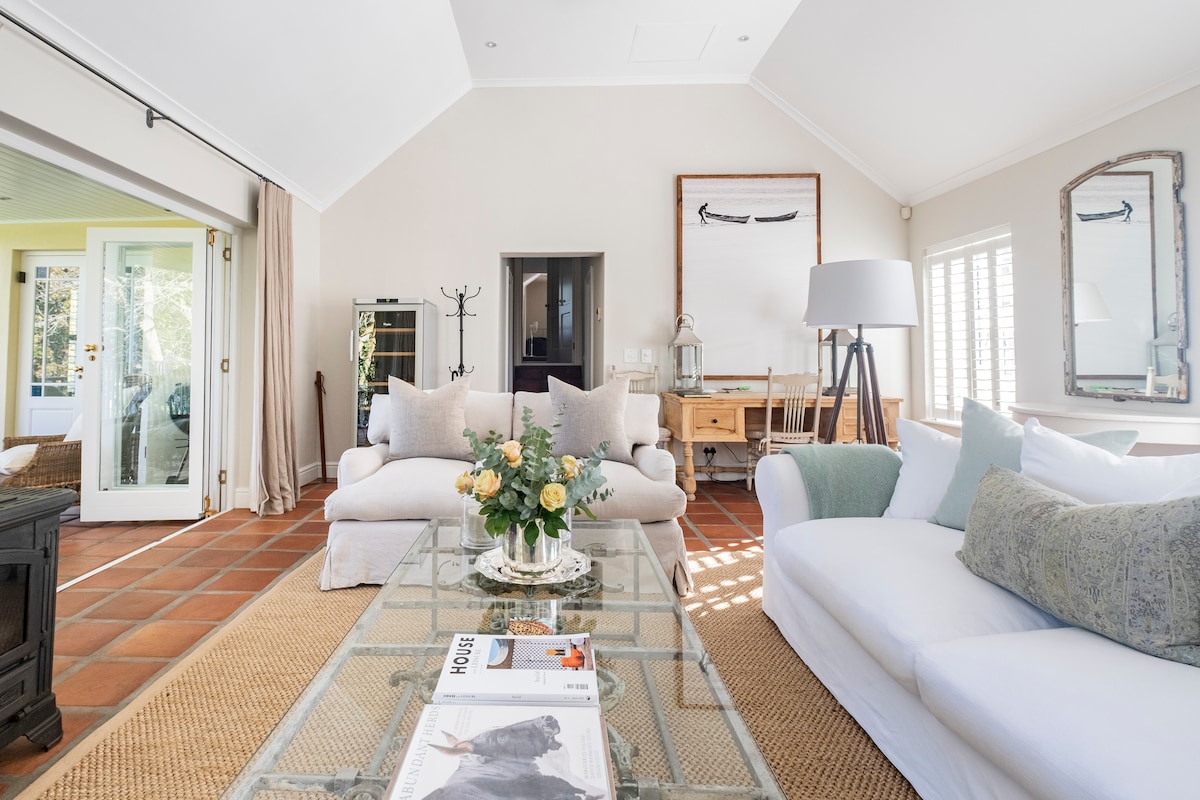 A bright living area features natural light streaming through large windows. Comfortable seating options include a light-colored couch and generous armchairs. A coffee table is adorned with a floral arrangement, while artwork and decorative mirrors enhance the space's style. Elegant patio doors lead to an outdoor terrace.