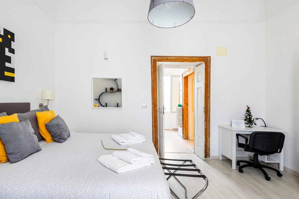 A bright bedroom features a spacious bed adorned with gray and yellow accents, alongside fresh white towels neatly arranged. A desk is positioned in one corner, and a doorway leads to additional living spaces, all illuminated by natural light from overhead fixtures.