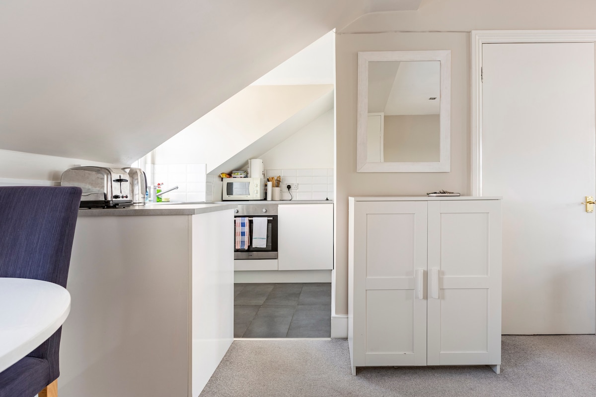 An airy studio flat features a compact kitchen with modern appliances, including a microwave and an electric kettle. A dining area is visible, with a round table and a single blue chair. Natural light illuminates the space from the angled ceiling.