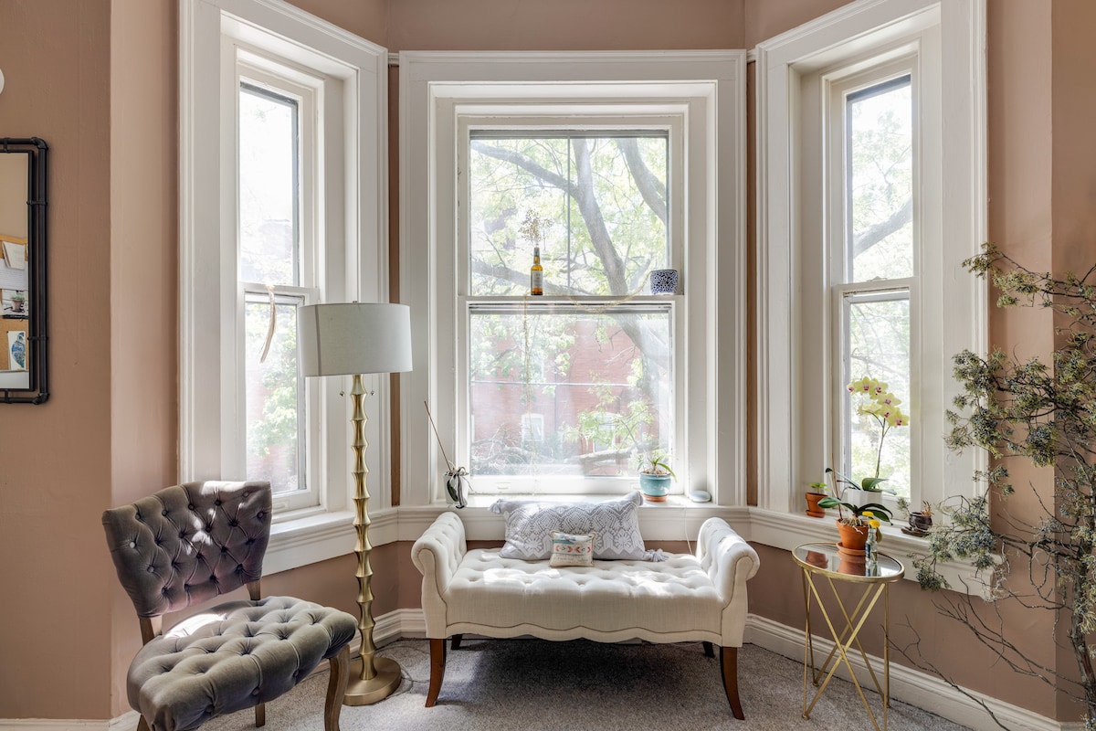 A charming seating area features a tufted white settee positioned between three tall windows, allowing natural light to fill the space. A small round table stands beside a sleek lamp, and greenery from potted plants is visible outside, creating a serene atmosphere.