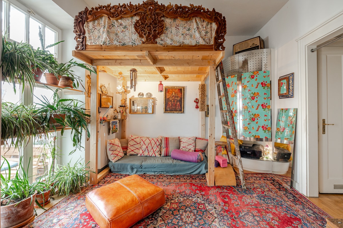 A unique living space features a lofted sleeping area above a cozy seating area, adorned with patterned cushions. Decorative plants line the windows, providing a fresh touch. An intricately carved headboard and vibrant area rug enhance the artistic character of the room.