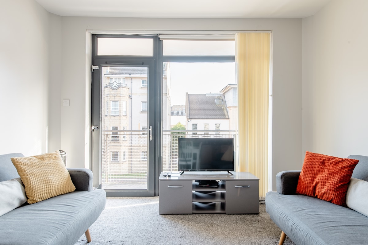 The living area features two comfortable gray sofa beds positioned across from a stylish media console with a flat-screen TV. Large glass doors open to a balcony, allowing natural light to fill the space, while sheer curtains softly diffuse the sunlight.