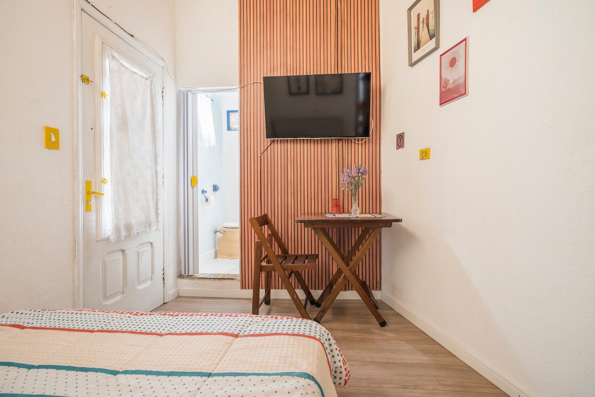 A cozy room features a comfortable bed with a colorful blanket. A wooden table and chair are positioned against a textured wall. A wall-mounted TV is visible, with a doorway leading to an adjacent bathroom in the background.