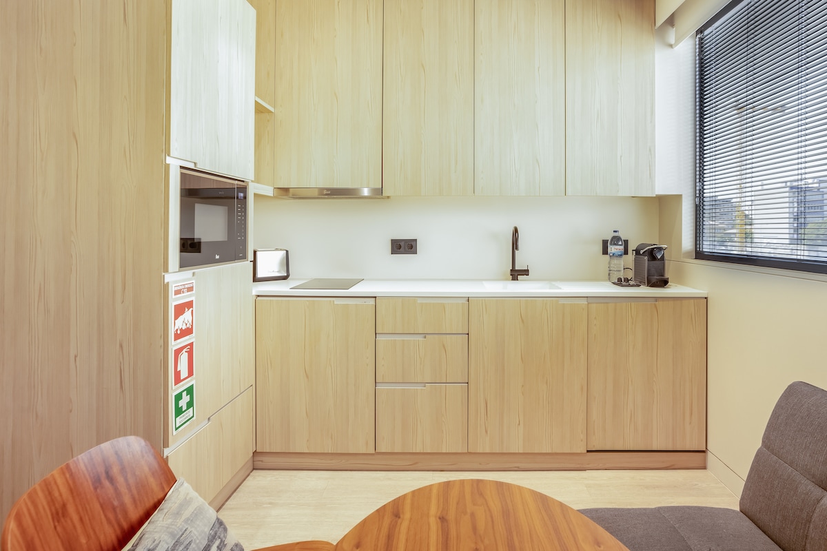 A modern kitchenette features light wood cabinetry and a muted countertop. Appliances include a microwave and a coffee machine, while a sleek faucet is mounted over the sink. A round wooden table and a gray seating area complete the inviting design.