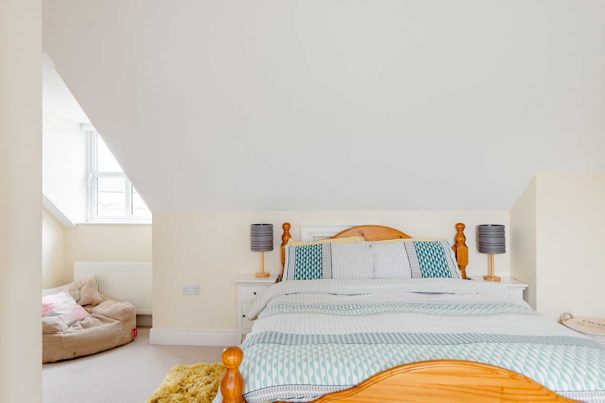 A spacious bedroom features a double bed with a patterned duvet cover. Two bedside lamps provide soft lighting. A cozy seating area with a plush large cushion is visible, positioned near a window that invites natural light. Neutral-colored walls create a calming atmosphere.
