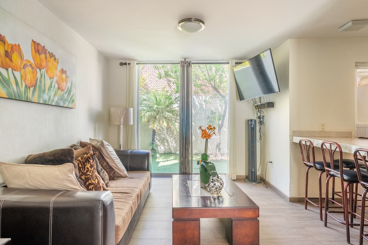 A well-lit living area features a comfortable sofa with decorative cushions and a wooden coffee table atop a light-colored floor. A large flat-screen TV is mounted on the wall, while sliding glass doors provide views of an outdoor space with greenery.