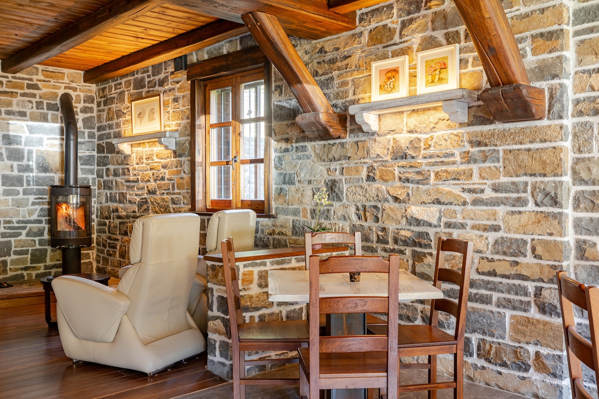 A rustic interior features a stone wall adorned with framed artwork. A wooden dining table, surrounded by matching chairs, is positioned next to an armchair. A modern stove stands in the corner, adding warmth to the space, which is illuminated by natural light through the window.