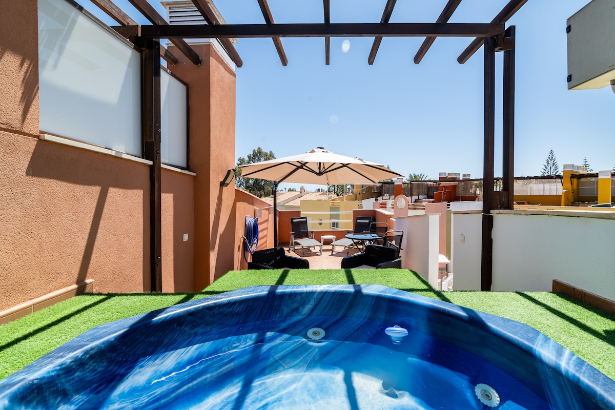 A private terrace features a jacuzzi surrounded by wooden decking and artificial grass. An umbrella provides shade over loungers positioned for relaxation. The space is framed by beams, allowing for a light, airy atmosphere with views of the surroundings.