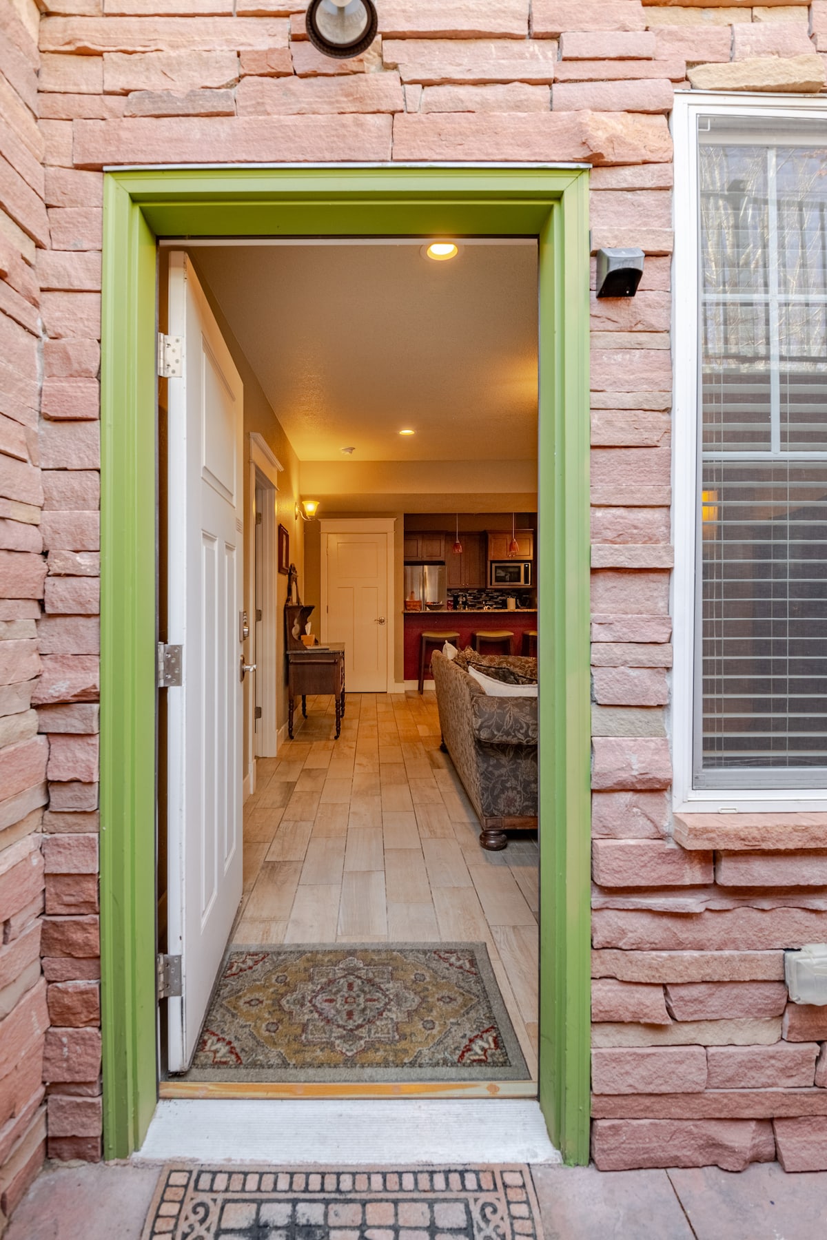A welcoming entrance is shown, framed by natural stone walls in warm hues. The open door reveals a glimpse of a spacious living area with wooden flooring and elegant decor. A patterned welcome mat lies at the doorstep, enhancing the inviting feel of the entrance.