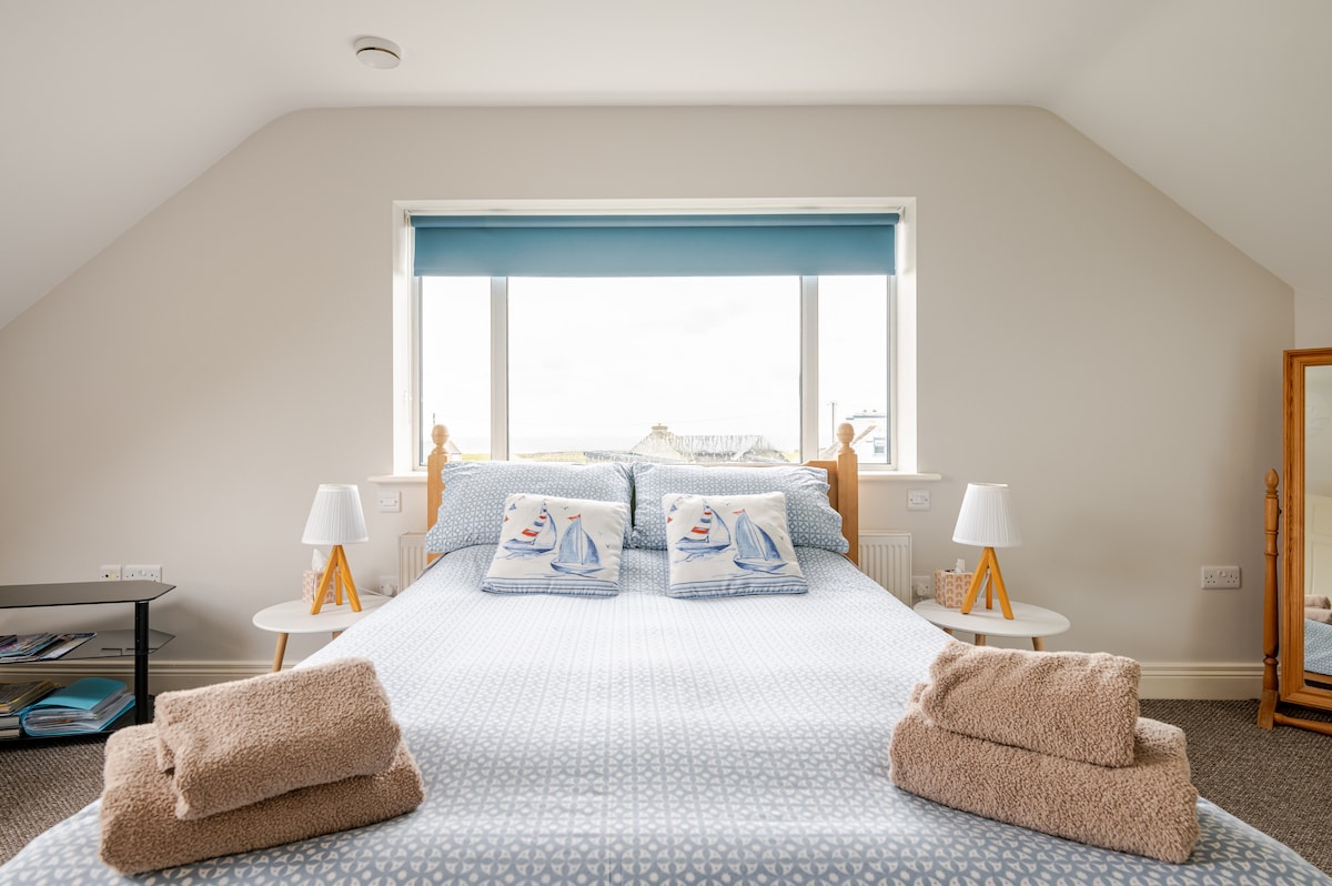 A spacious bed is positioned in front of a large window, providing natural light. Soft blue and white bedding is complemented by decorative pillows. Two bedside tables with lamps frame each side of the bed, while a full-length mirror and a small table are visible nearby.