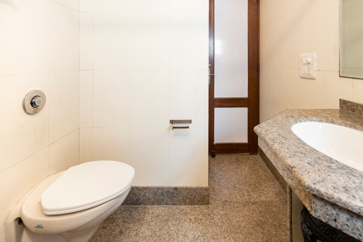 A clean and well-maintained bathroom features a modern toilet and a granite countertop with a sink. Neutral wall tiles provide a simple backdrop, and a wooden door is visible, allowing for privacy.