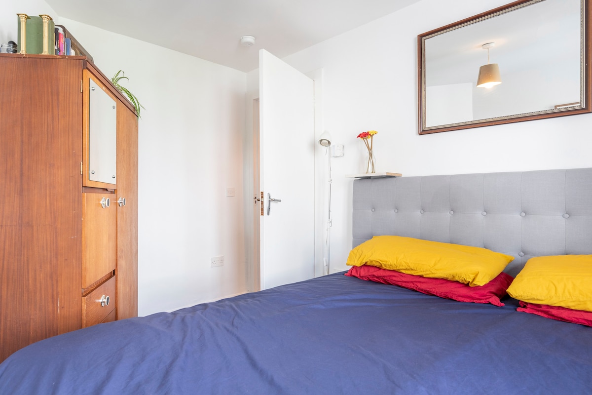 A bright bedroom is presented, featuring a spacious bed with layered yellow and red pillows. A wooden wardrobe stands beside the door, contributing to the room's simplicity. A mirror reflects the soft light from an overhead lamp, creating a welcoming environment.