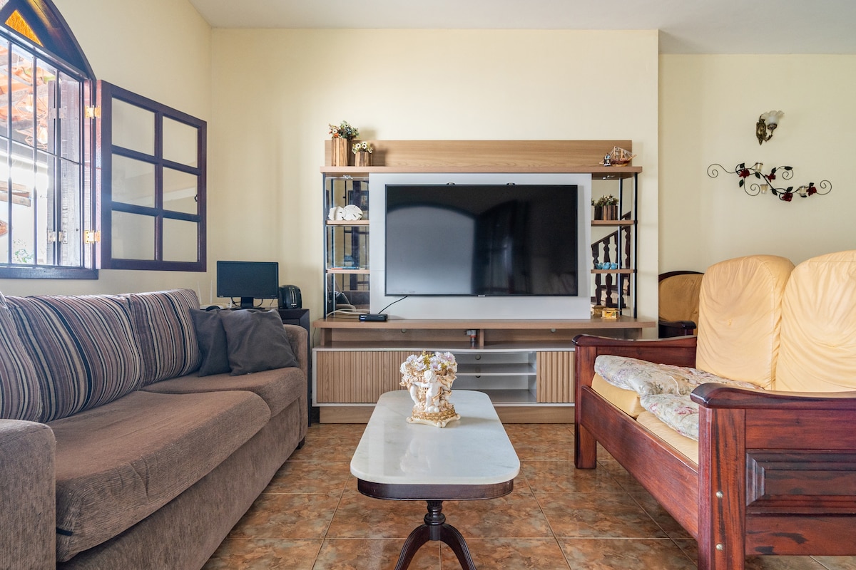 A spacious living area features a large flat-screen TV mounted on a wooden cabinet. Two comfortable armchairs are positioned opposite a soft sectional sofa adorned with patterned cushions. A small coffee table sits at the center on a tiled floor under natural light from adjacent windows.