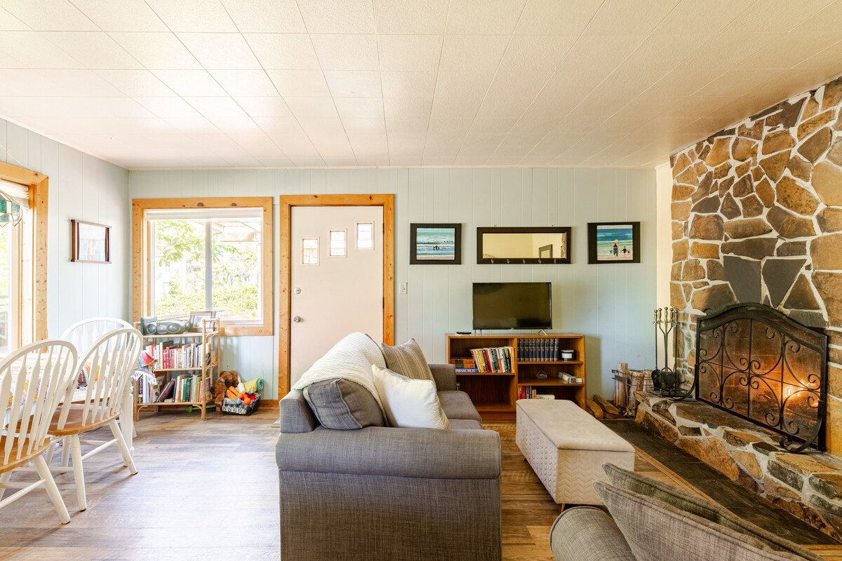 A cozy living area features a large, inviting sofa and an ottoman, with natural light streaming in through the door and windows. A stone fireplace adds character, while a modest entertainment center with a TV and bookshelves completes the space. White dining chairs surround a wooden table.