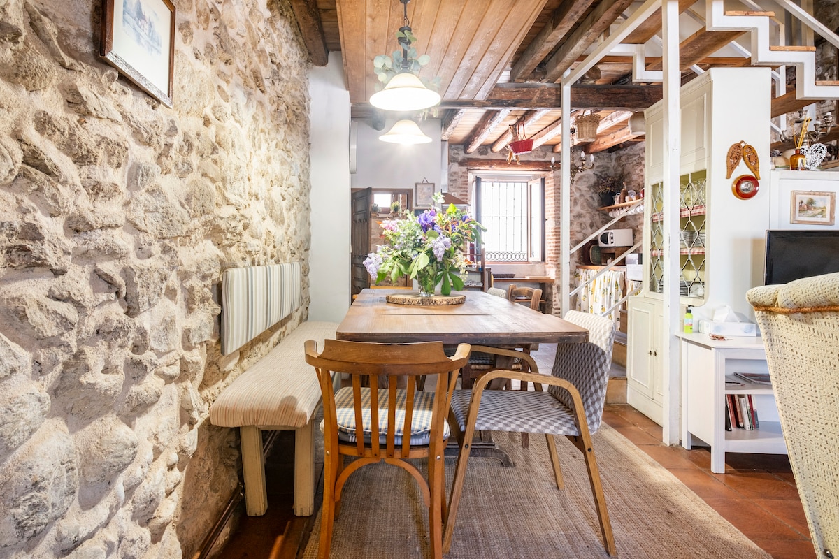 A rustic dining area features a wooden table surrounded by various chairs, complemented by a bouquet of fresh flowers. Exposed stone walls and wooden beams create a warm ambiance, while a staircase is visible in the background, leading to additional living spaces.