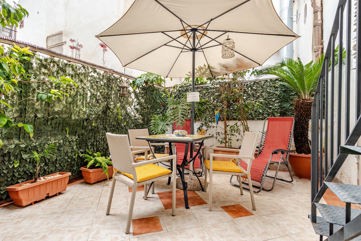 A tranquil outdoor terrace features a shaded table with chairs and lounge seating. Lush greenery surrounds the space, enhancing the natural atmosphere. A large umbrella provides shade, while potted plants add color and warmth to the area.
