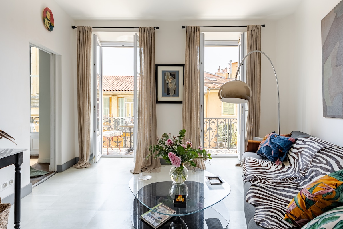 A light-filled living area features an airy atmosphere, with large open windows allowing sunlight to enter. A patterned sofa is paired with a glass coffee table adorned with decorative items. A modern arc lamp adds style, and a vibrant artwork hangs on the wall.