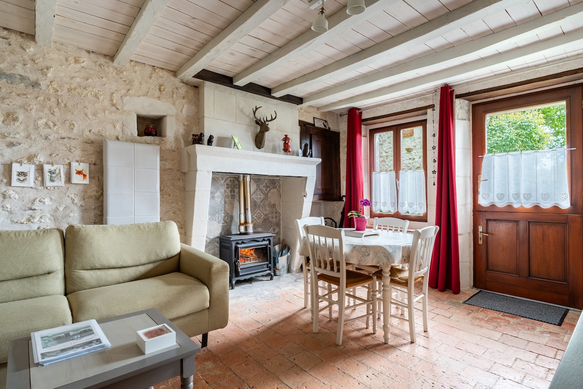A warm living area features a stone fireplace with a wooden mantel, complemented by a cozy sofa and an inviting dining table set for four. Natural light enters through double doors adorned with lace curtains, enhancing the rustic charm of the space.