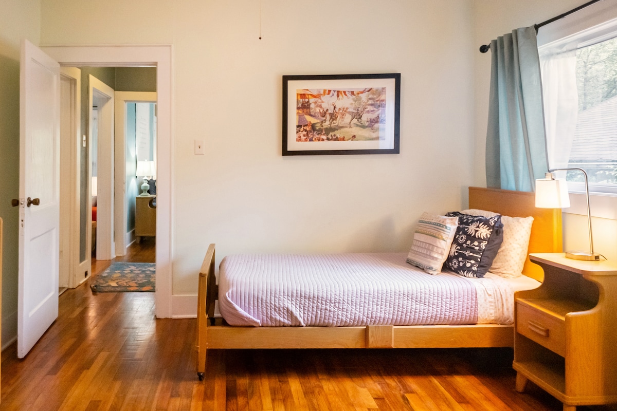 A cozy bedroom features a single wooden bed with a fitted sheet and decorative pillows. A bedside table holds a lamp, providing light. A piece of art hangs above the bed, and a large window reveals soft natural light. The room connects to a hallway leading to other areas.