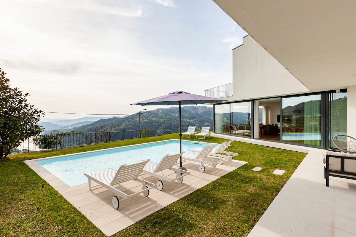 An outdoor space features a private pool surrounded by a well-maintained lawn. Sun loungers are arranged under a large umbrella, offering shade. Glass doors provide a view of the interior, while a gentle slope of hills can be seen in the background.