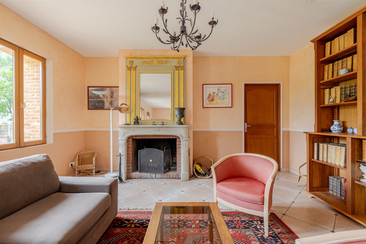 A well-lit living area features a comfortable gray sofa and a pink chair, arranged around a wooden coffee table. A classic fireplace with a decorative mirror above serves as a focal point, while built-in shelves display books and decorative items. Large windows offer natural light.