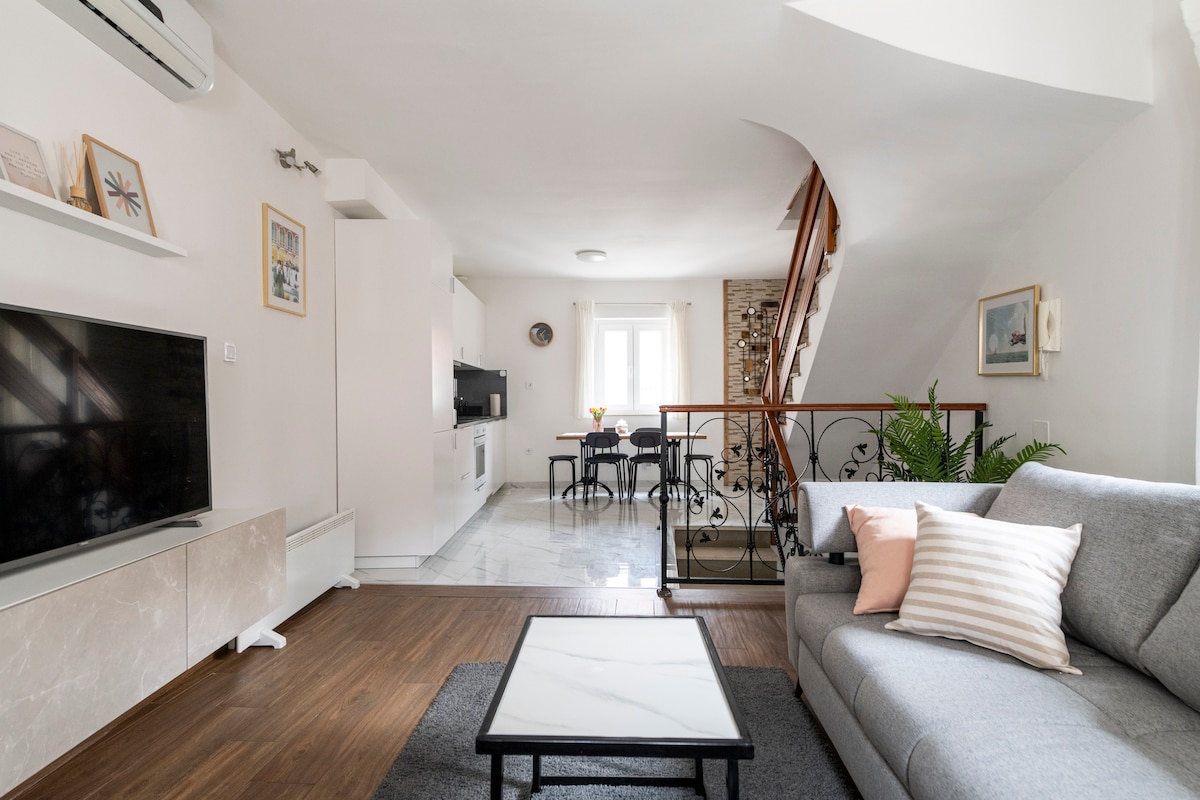 A living area features a light gray sofa with decorative cushions, positioned near a large TV and a sleek media console. A dining table with chairs is visible in the background, adjacent to a window that allows natural light to fill the room. Stairs lead to the upper floor.