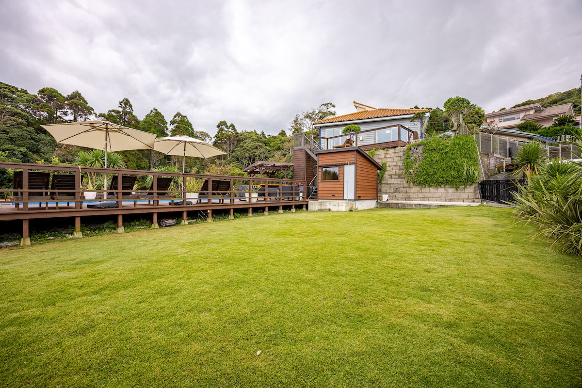 A well-maintained green lawn encircles a wooden deck and a cozy cabin, suggesting an inviting outdoor space. Several patio umbrellas provide shade over the deck, which is positioned against a backdrop of lush vegetation and a distant ocean-view residence.