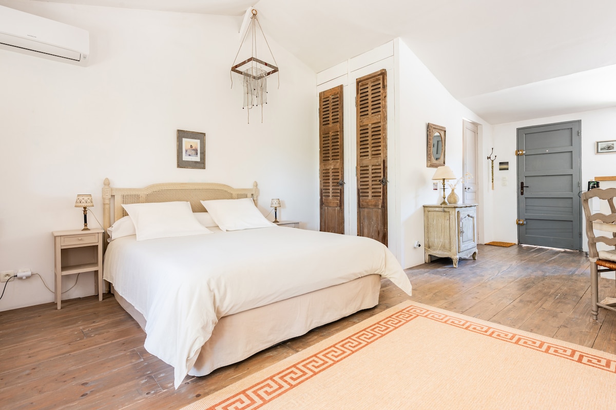 A spacious bedroom features a large, comfortable bed with two plush pillows. Light-colored walls are complemented by a wooden floor and subtle area rug. Vintage storage units are visible, along with bedside tables and lamps, creating a calm and organized atmosphere.