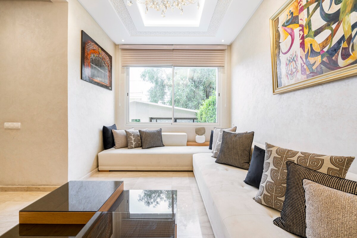 A serene living room features a comfortable white sectional sofa adorned with decorative pillows in various textures. A wooden coffee table is positioned in front of the seating area. Natural light enters through a large window, complemented by modern artwork on the walls.
