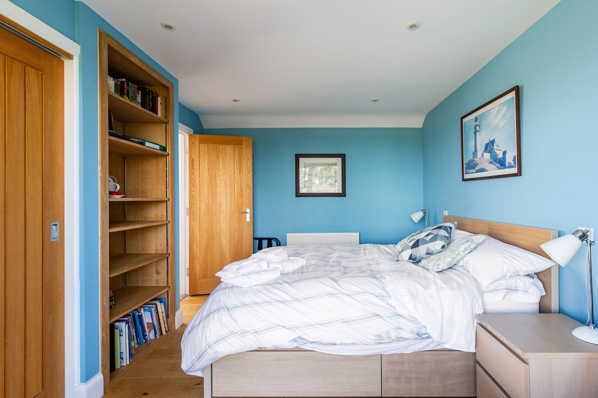 A light-filled bedroom features blue walls and wooden accents. A spacious bed is adorned with soft linens, complemented by bedside lamps. A wooden bookshelf holds books, while an art piece depicting a lighthouse hangs on the wall. A door leads to an adjacent room.