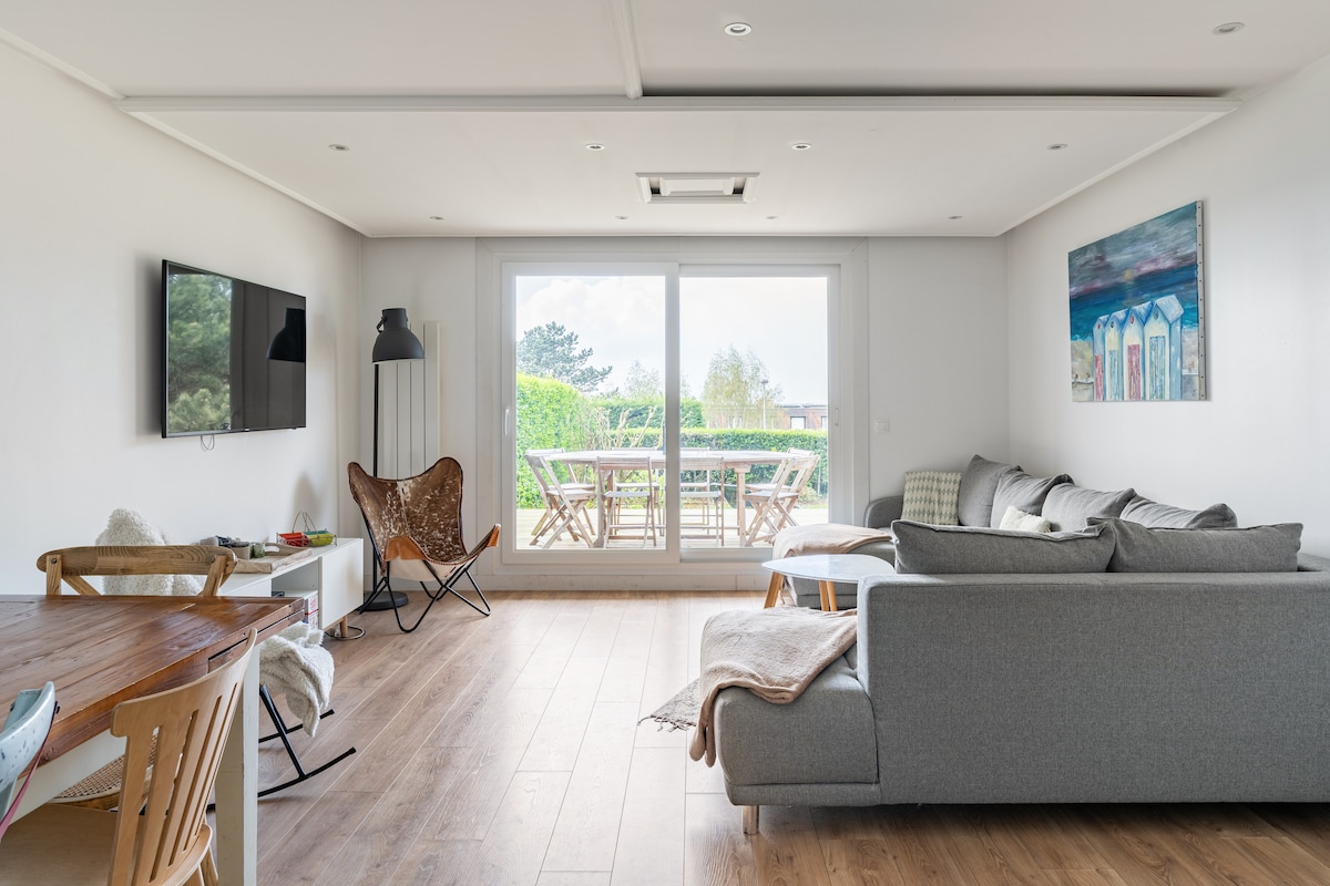 A spacious living area features a large grey sofa and a stylish table with wooden chairs. Floor-to-ceiling glass doors offer views of the outdoor terrace and garden, creating a bright and open atmosphere. A wall-mounted television and a piece of artwork add to the decor.